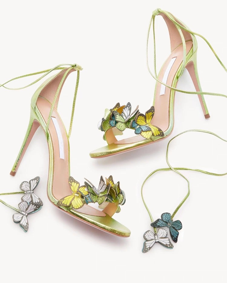 Gold stiletto high heels with butterfly and flower appliques, ankle wrap straps, open toe design.