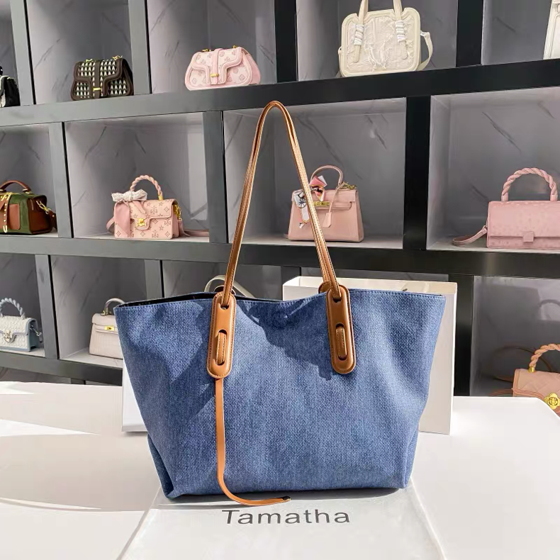 Blue tote bag with brown straps on a white surface with 'Tamatha' branding, shelves with more bags in the background.
