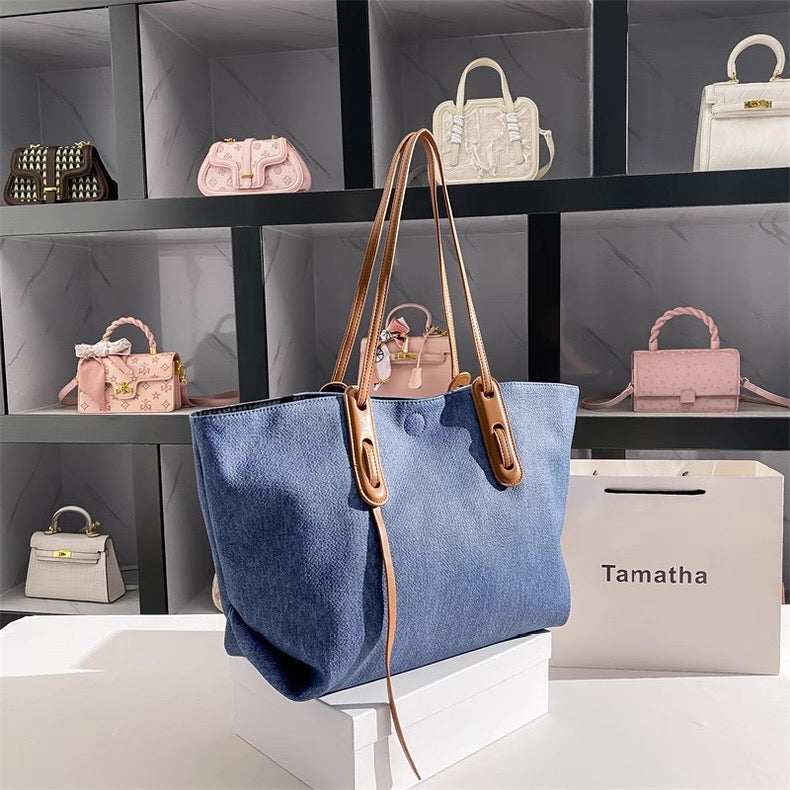 Blue tote bag with brown straps on a white platform, with shelves of handbags in the background.