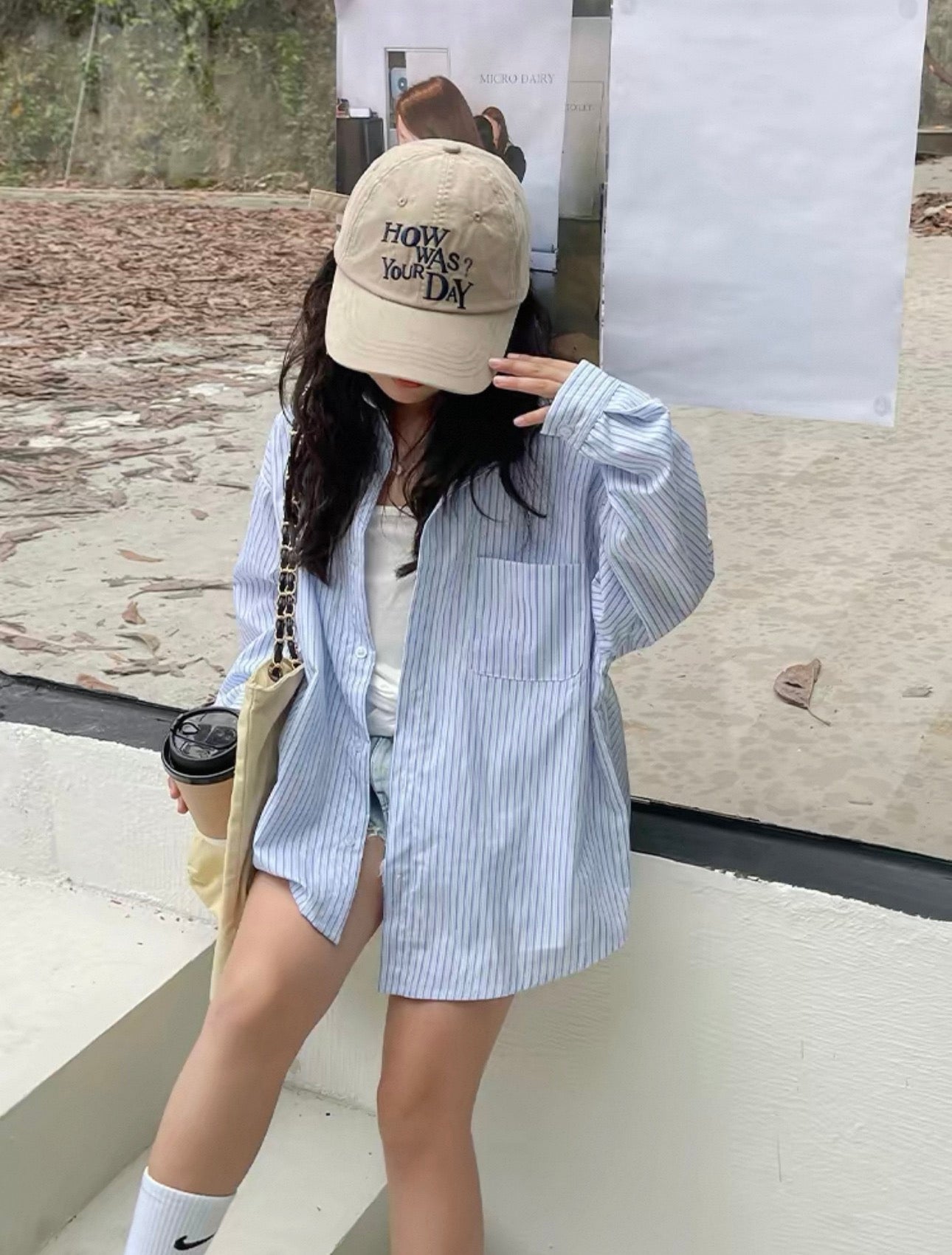 Person wearing a beige cap with text, blue striped shirt, and shorts, holding a coffee cup.