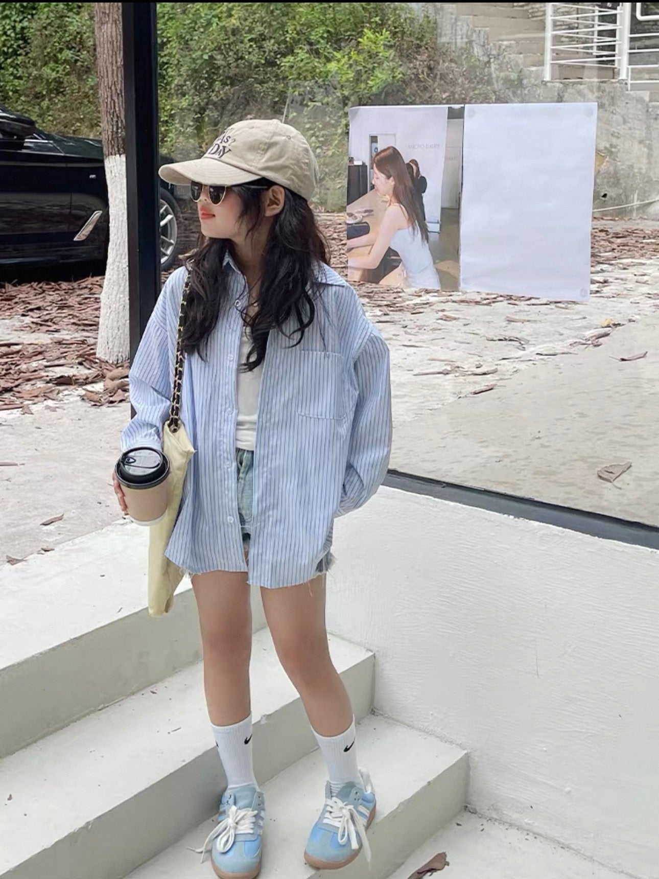 Woman standing on steps wearing a light blue striped shirt, denim shorts, and white sneakers.