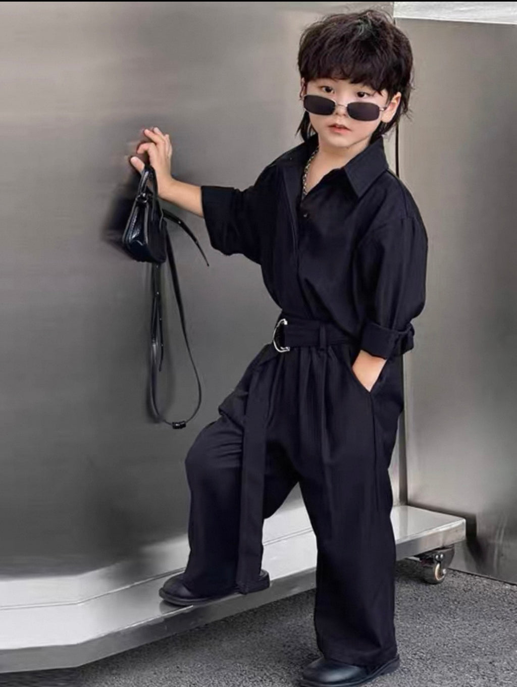 Child wearing a black outfit with sunglasses, sitting on a bench against a gray wall.