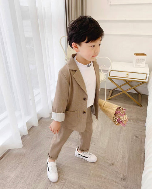 Child in a beige suit standing in a room with white curtains and a small table.