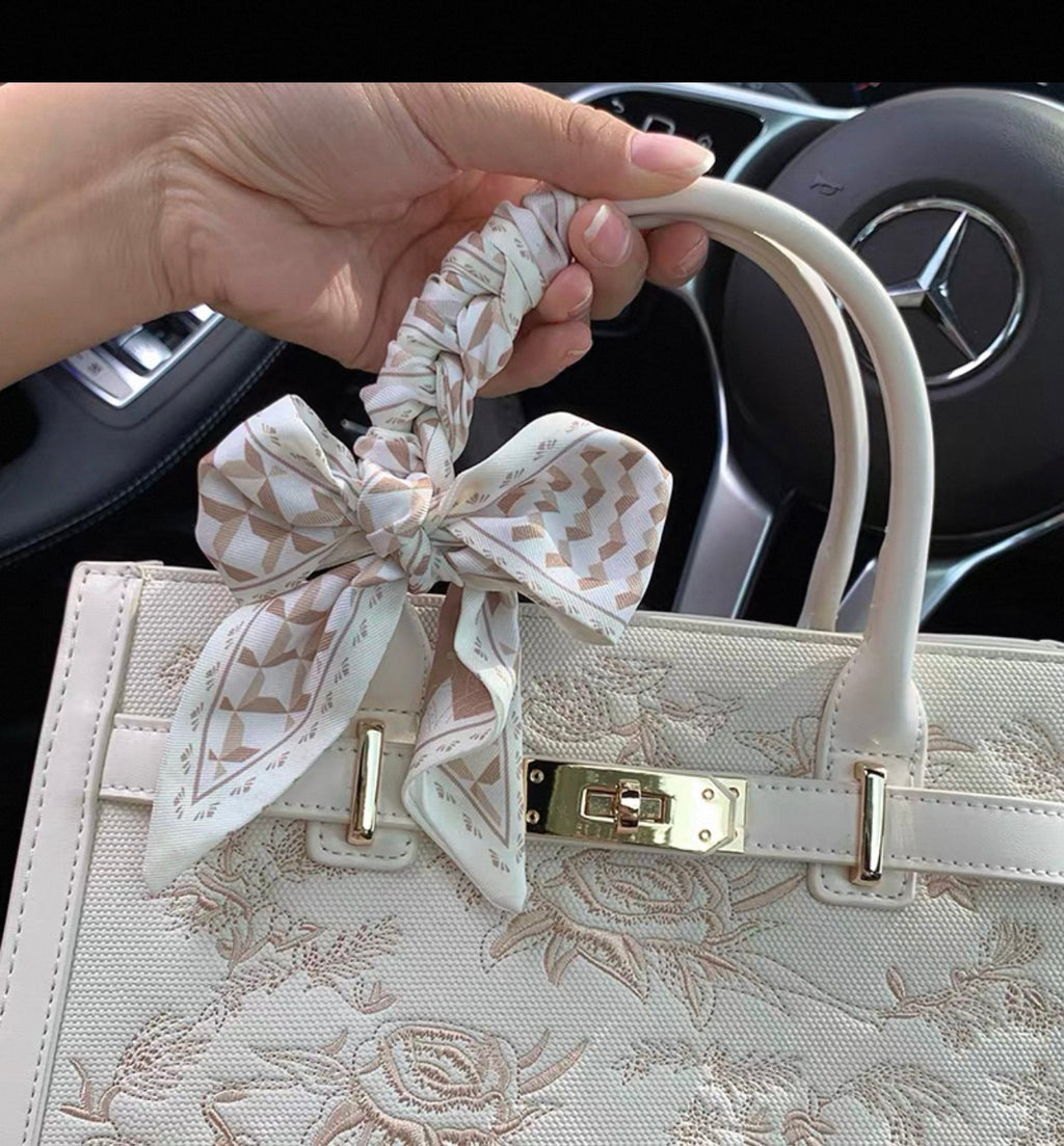 Cream-colored rectangular handbag with floral embroidery, gold-tone clasp, and a patterned fabric bow on one handle.
