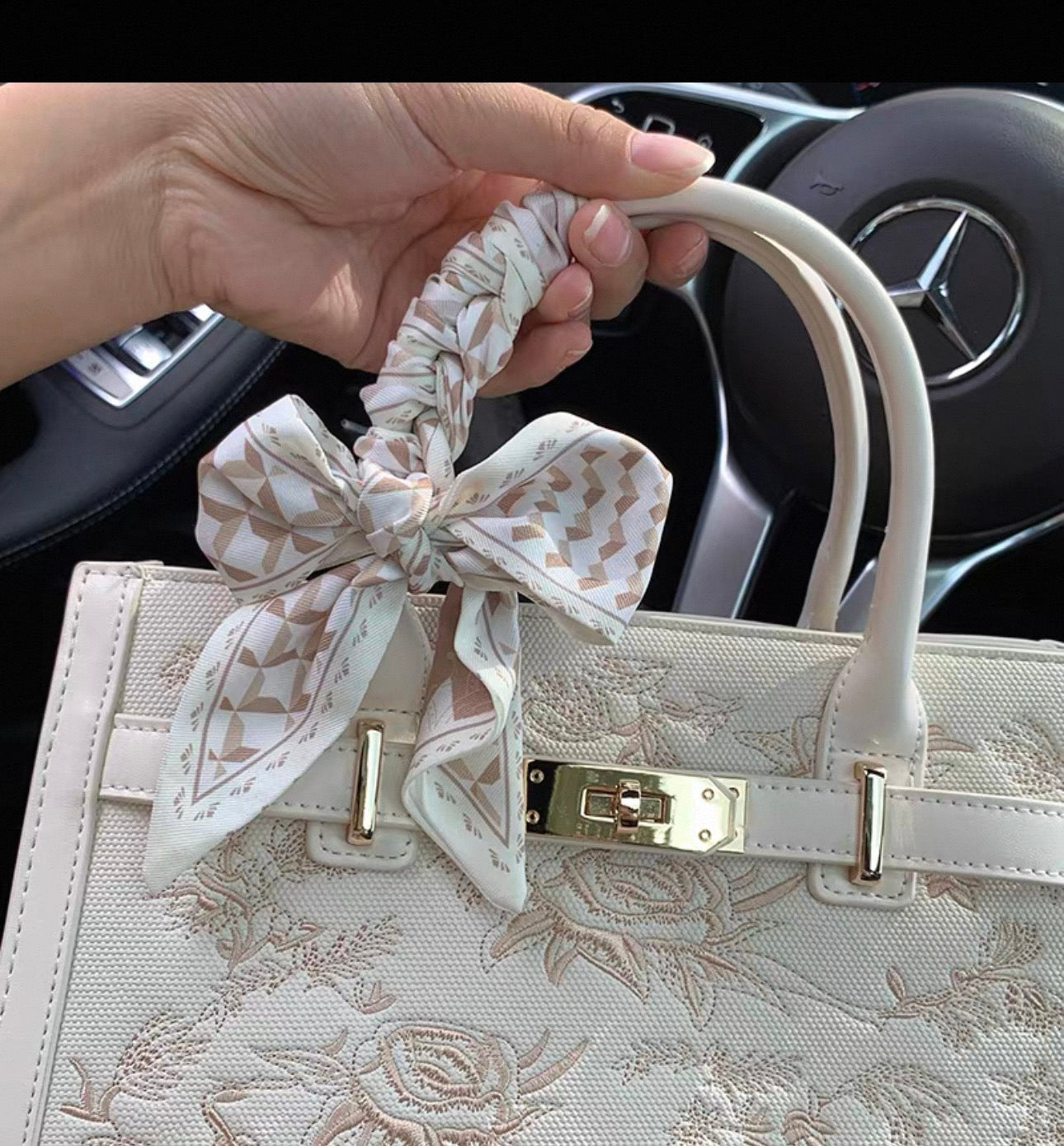 Cream-colored rectangular handbag with floral embroidery, gold-tone clasp, and a patterned fabric bow on one handle.