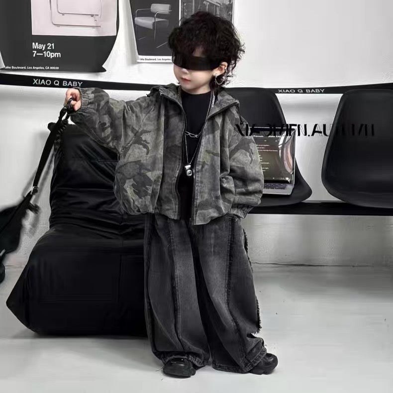 Person wearing a camouflage jacket and dark pants in a minimalistic setting with a chair and table in the background.