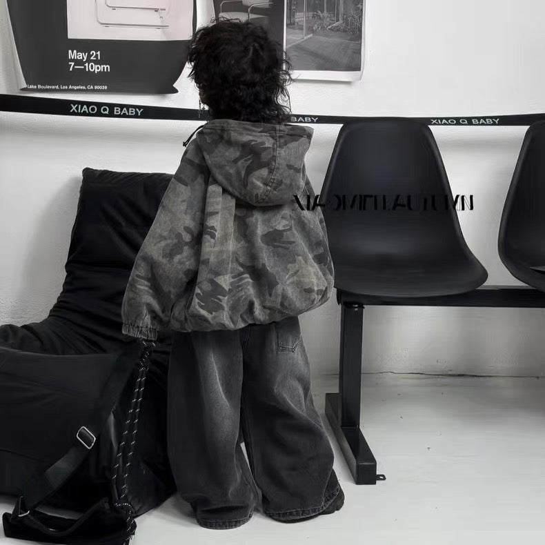 Person wearing a camouflage jacket and dark pants sitting on a black chair in a minimalistic room.