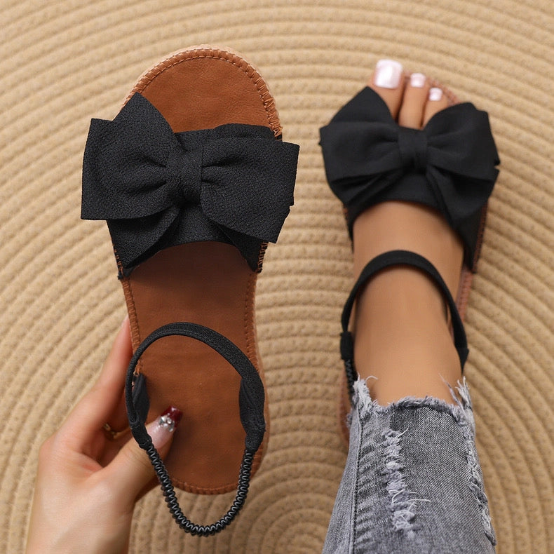 Black sandals with large bows worn on a textured beige surface