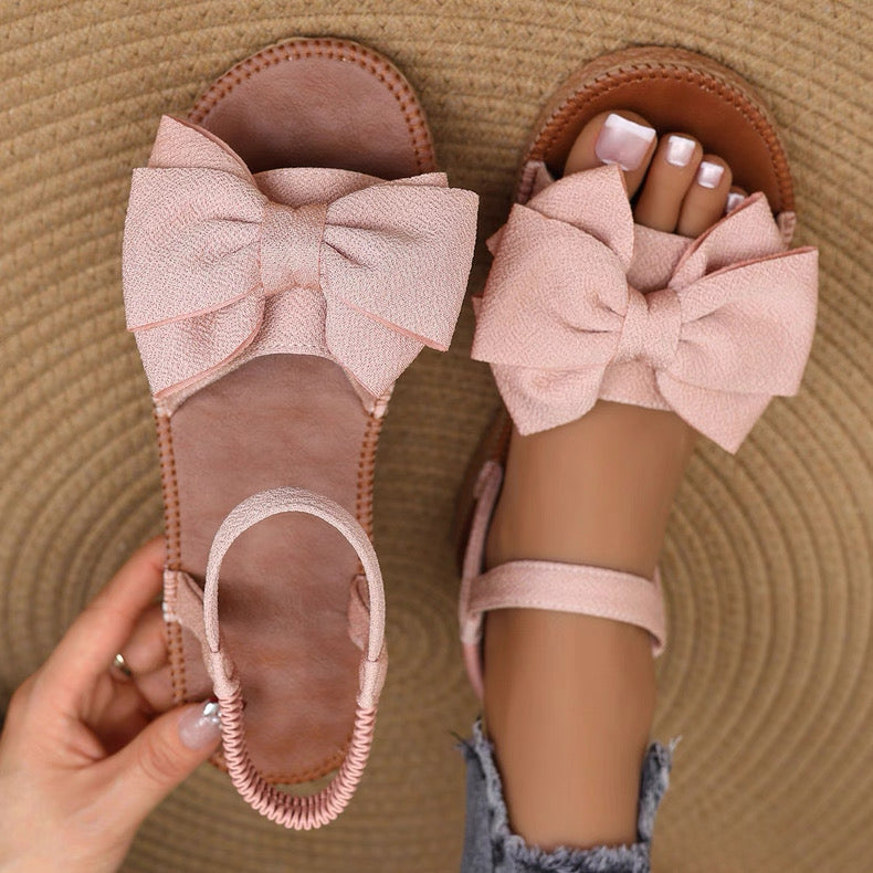 Pink sandals with large bows worn by a person on a textured brown surface