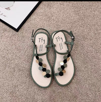 Pair of green sandals with pom-pom details on a carpeted floor.
