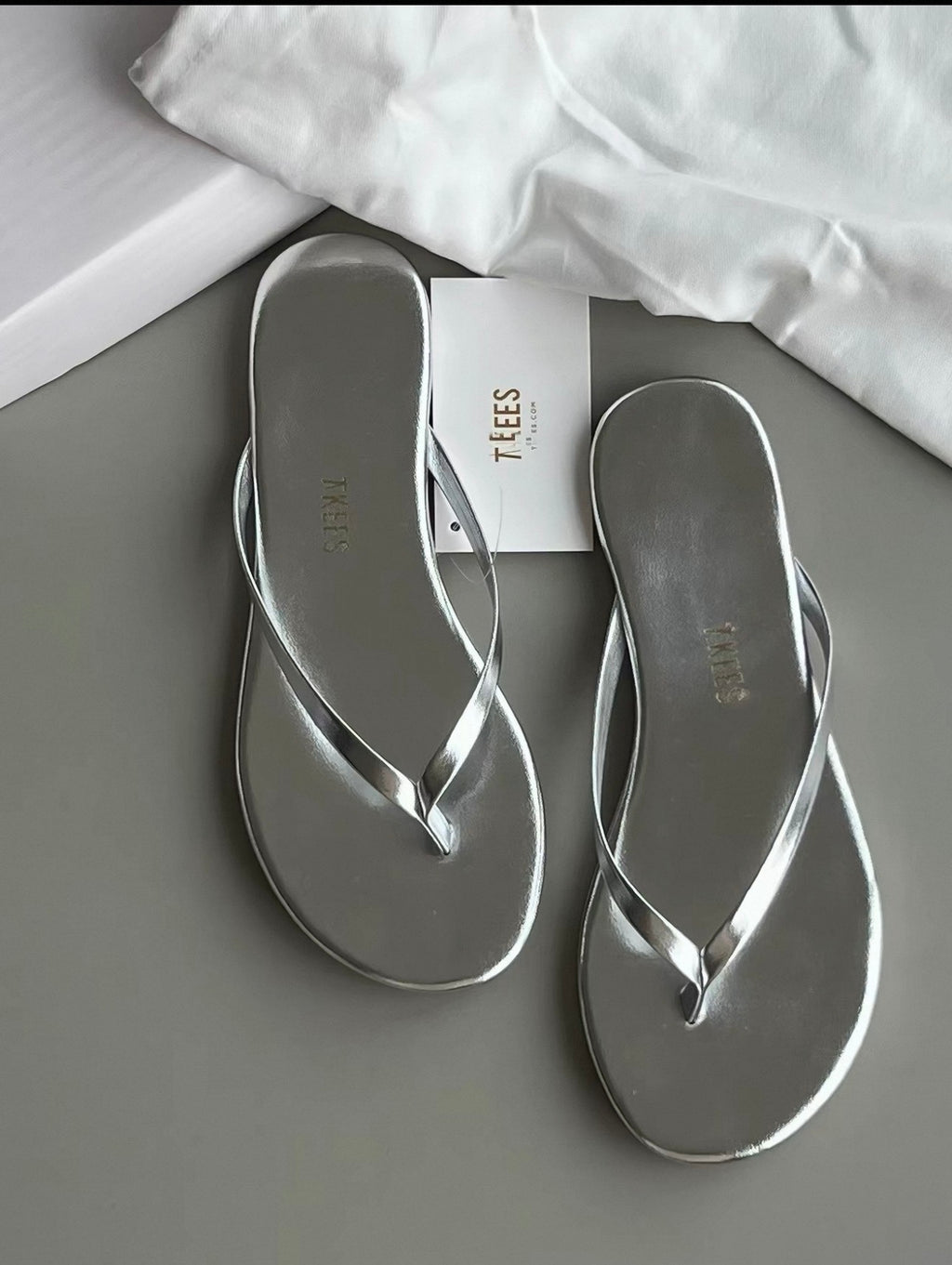 Silver flip-flops on a gray surface with a white cloth and 'TEES' box in the background.
