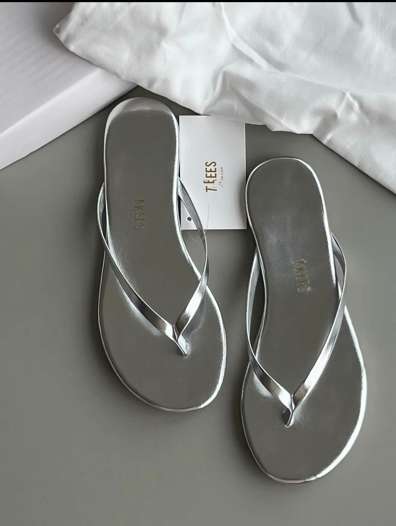 Silver flip-flops on a gray surface with a white cloth and 'TEES' box in the background.