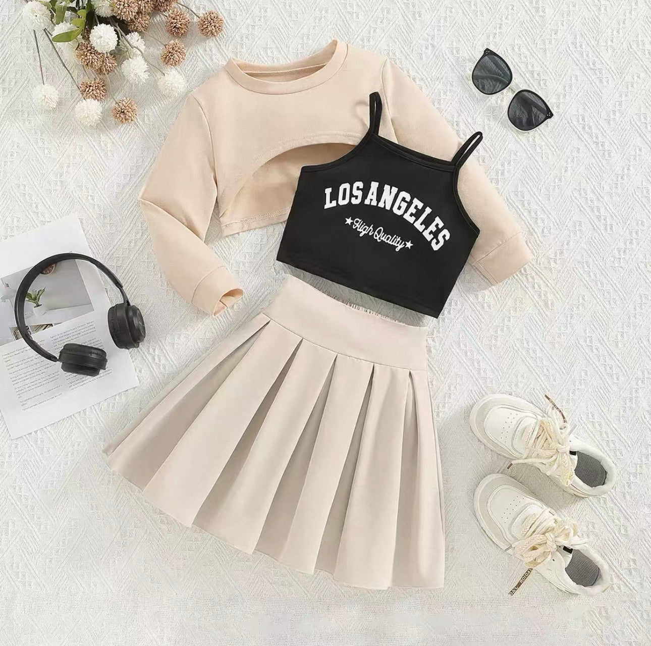 Beige long-sleeve top, black 'Los Angeles' crop top, beige pleated skirt, white sneakers, sunglasses, and headphones on a textured white surface.