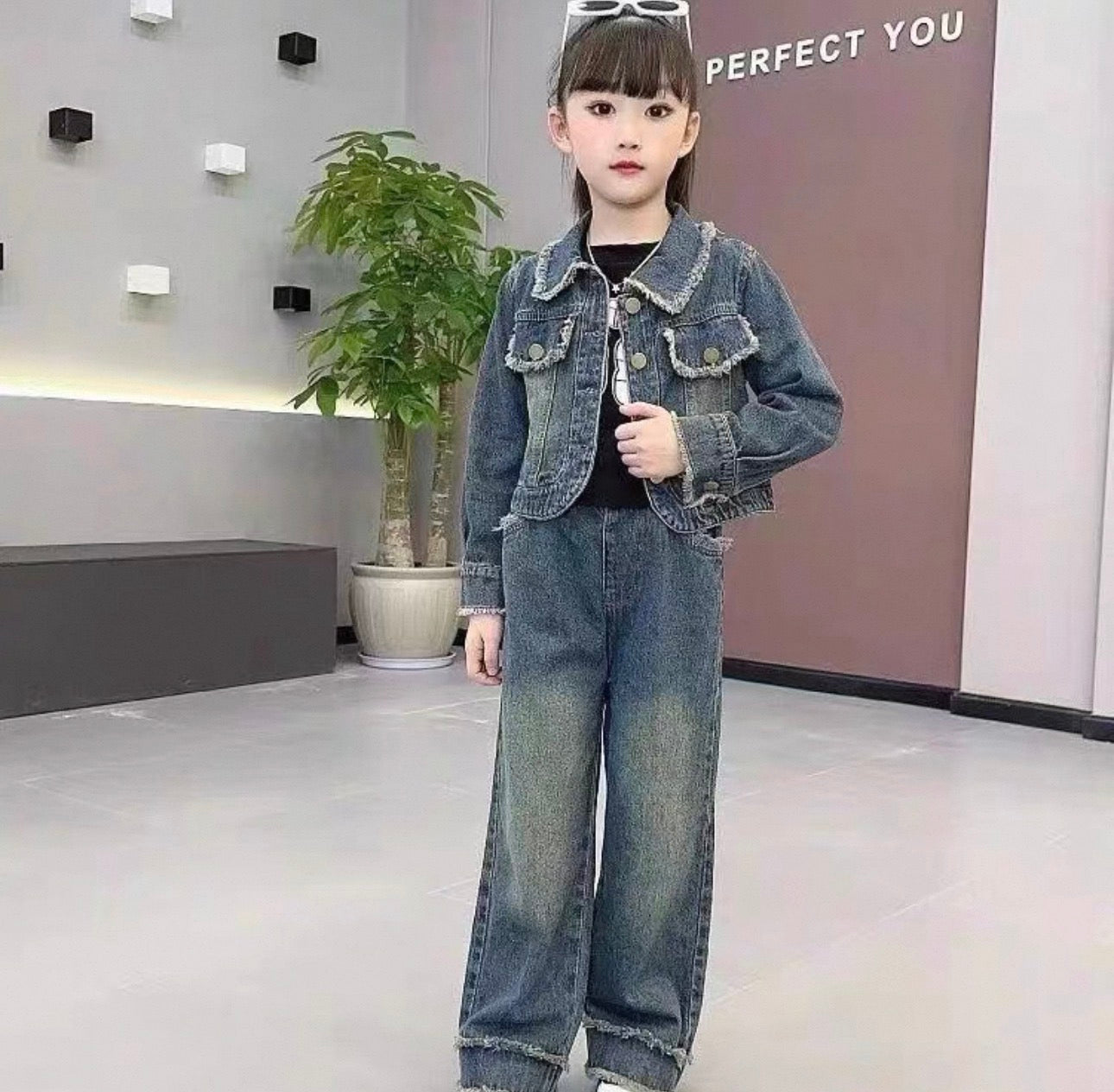 Child wearing a denim jacket and jeans in an indoor setting with a plant and wall text.