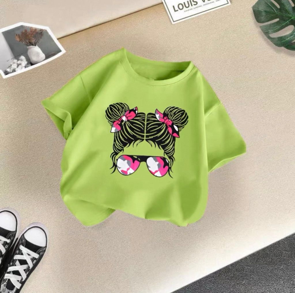 Green t-shirt with a graphic design of hair buns and sunglasses on a beige surface.