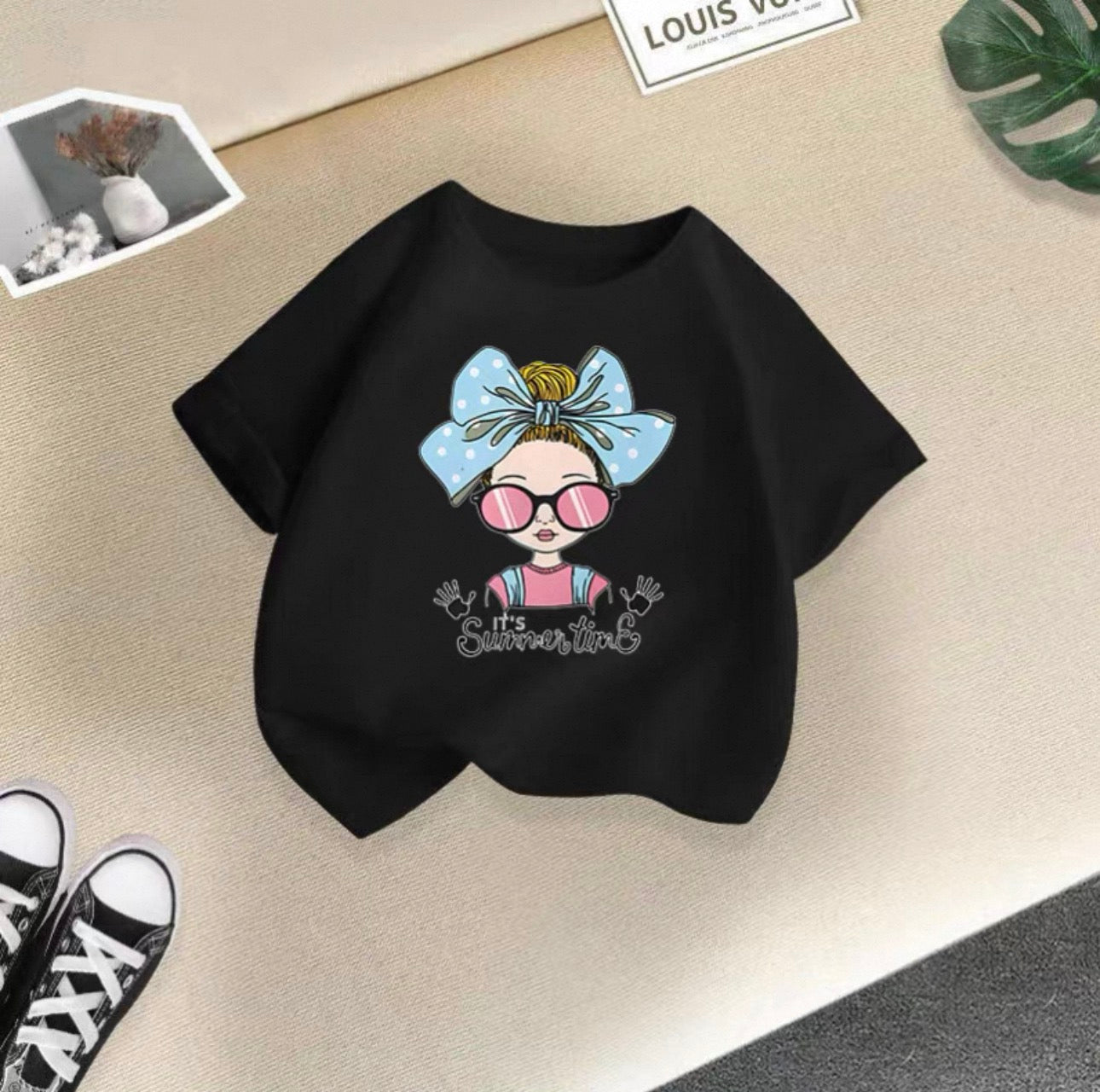 Black t-shirt with a cartoon design of a girl with a bow and sunglasses on a beige surface.