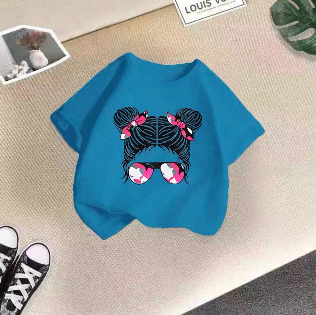 Blue t-shirt with a graphic design of hair buns and sunglasses on a beige surface.