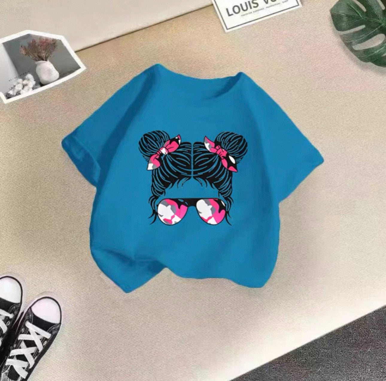 Blue t-shirt with a graphic design of hair buns and sunglasses on a beige surface.
