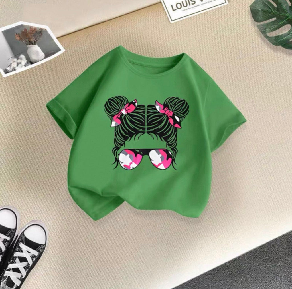 Green t-shirt with a graphic design of hair buns and sunglasses on a beige surface.