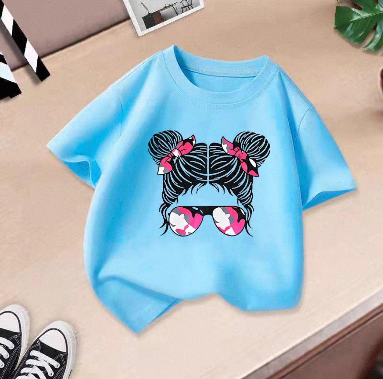 Light blue t-shirt with a graphic design of two buns with bows and sunglasses on a beige surface.