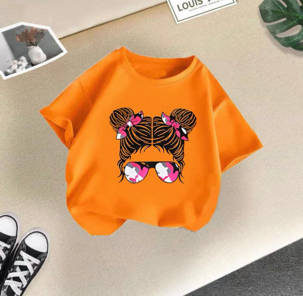Orange t-shirt with a graphic design of hair buns and sunglasses on a beige surface.