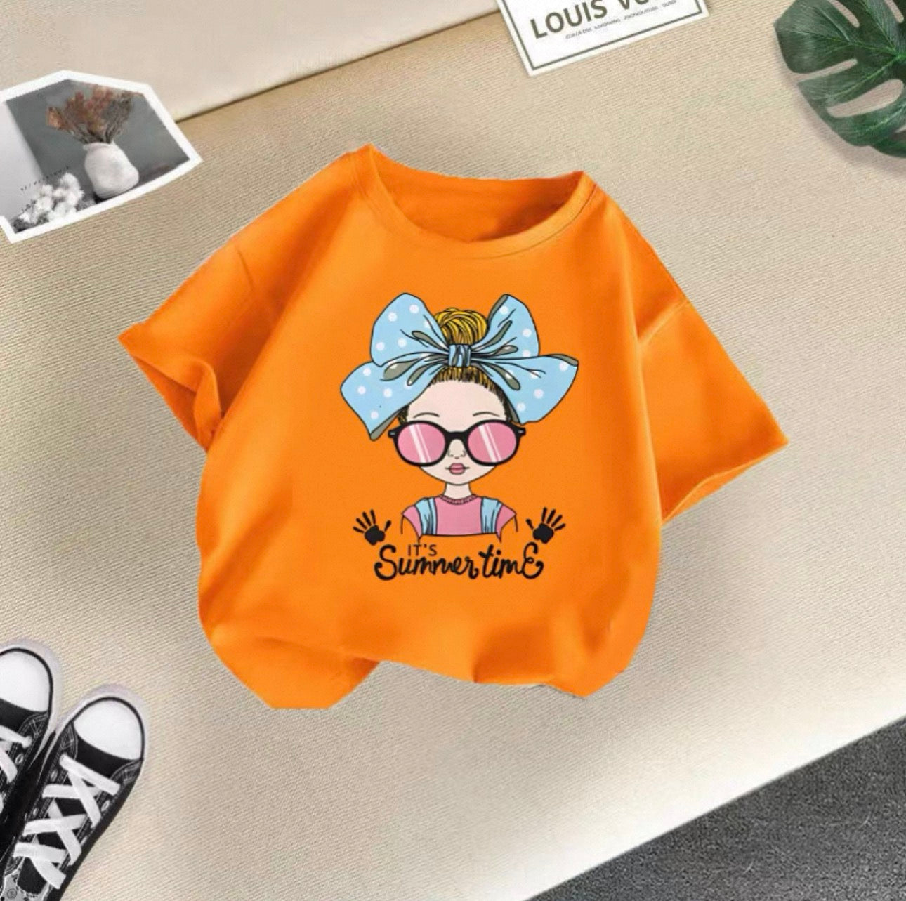 Orange t-shirt with a cartoon character and 'It's Summertime' text on a beige surface.