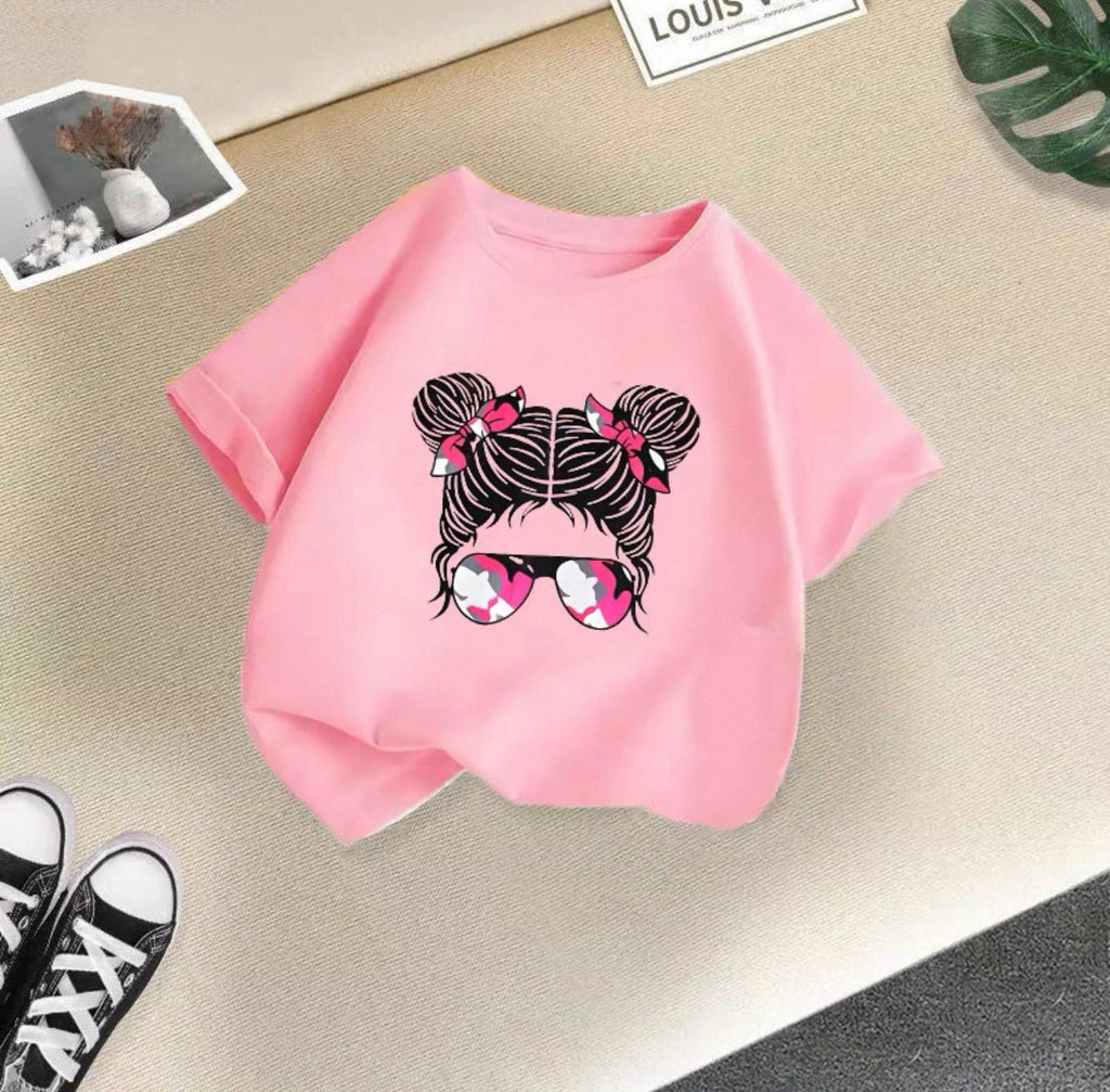 Pink t-shirt with a graphic design of a girl with braids and sunglasses on a beige surface.