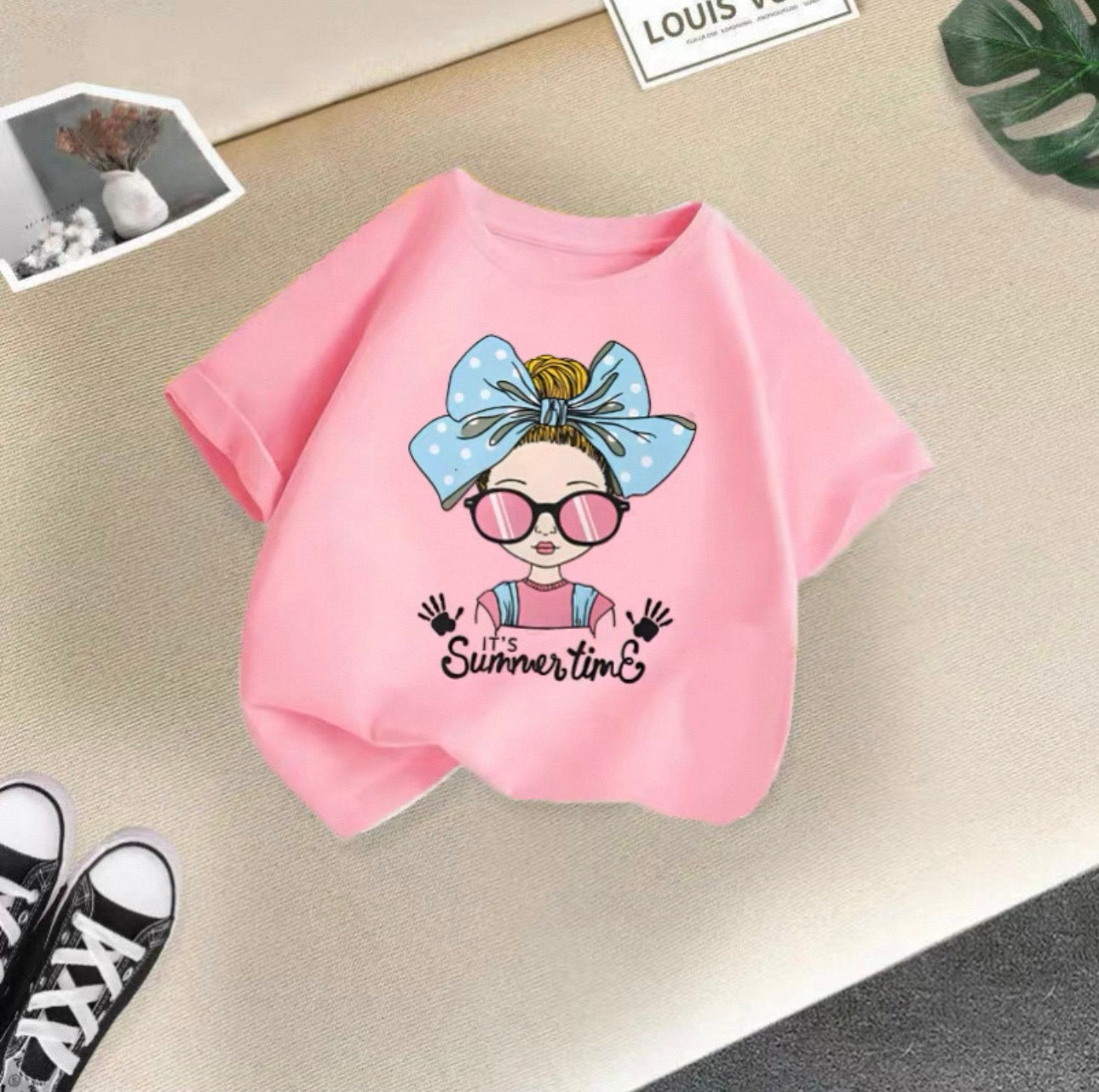 Pink t-shirt with a graphic design of a girl with a bow and sunglasses on a beige surface.
