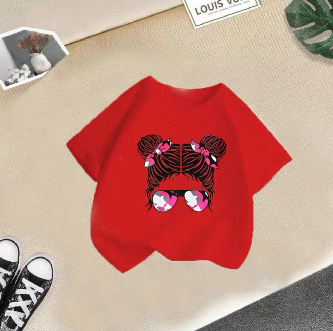 Red t-shirt with a graphic design of hair and sunglasses on a beige surface.