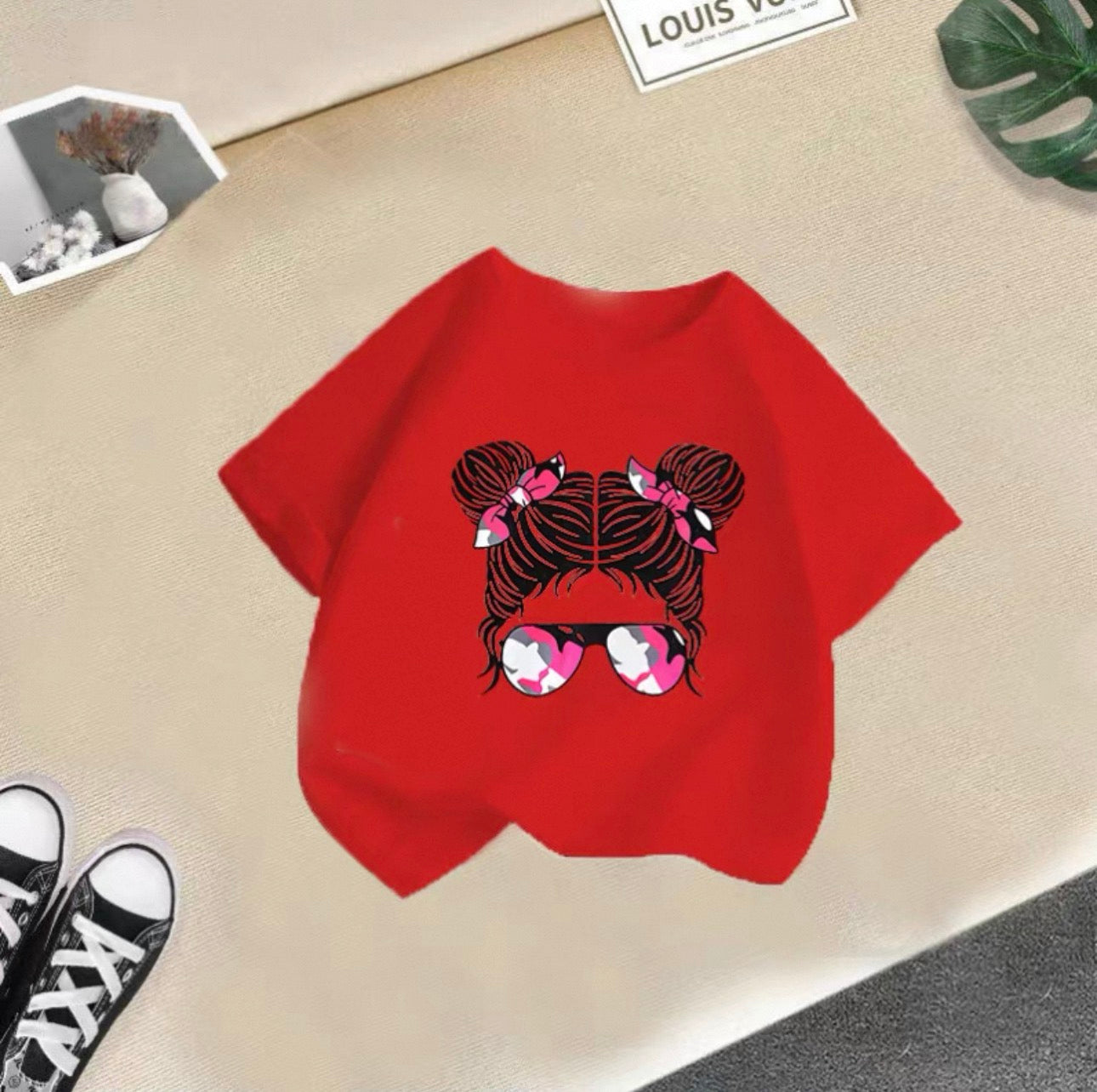 Red t-shirt with a graphic design of braids and sunglasses on a beige surface.