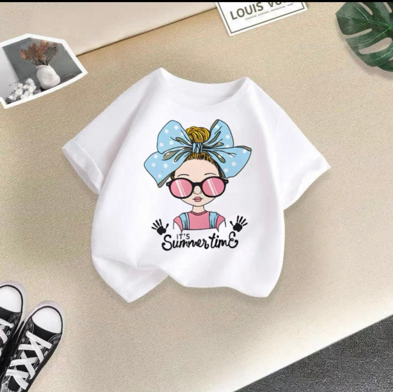 White t-shirt with a graphic design of a girl with a bow and sunglasses, text 'It's Summertime', on a beige surface.