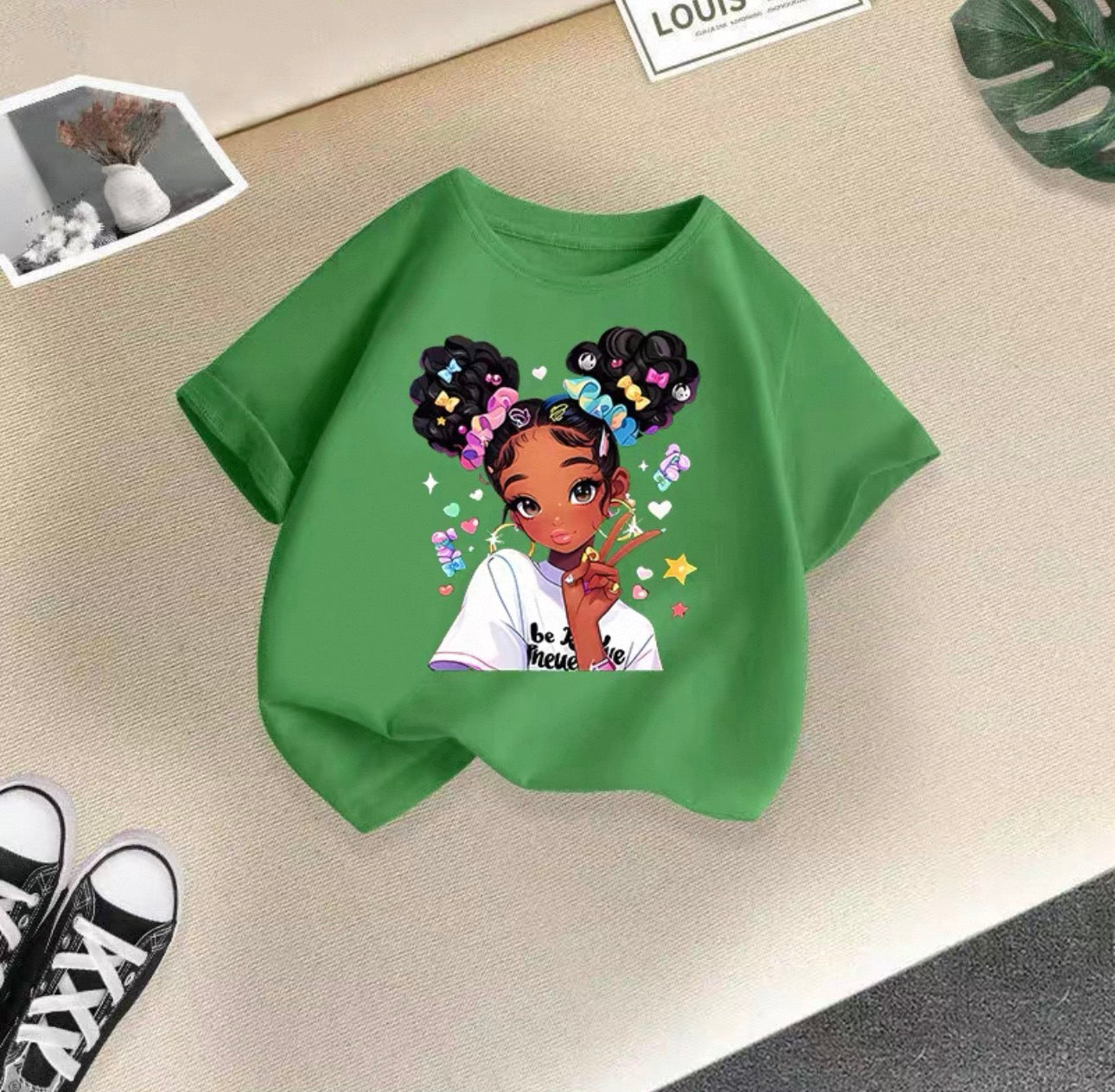 Green t-shirt with a graphic of a girl with braids and colorful letters on a beige surface.