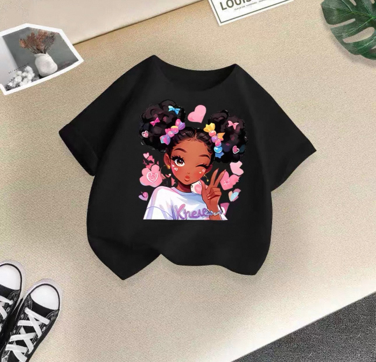 Black t-shirt with a colorful graphic of a girl with braids and flowers on a beige surface.