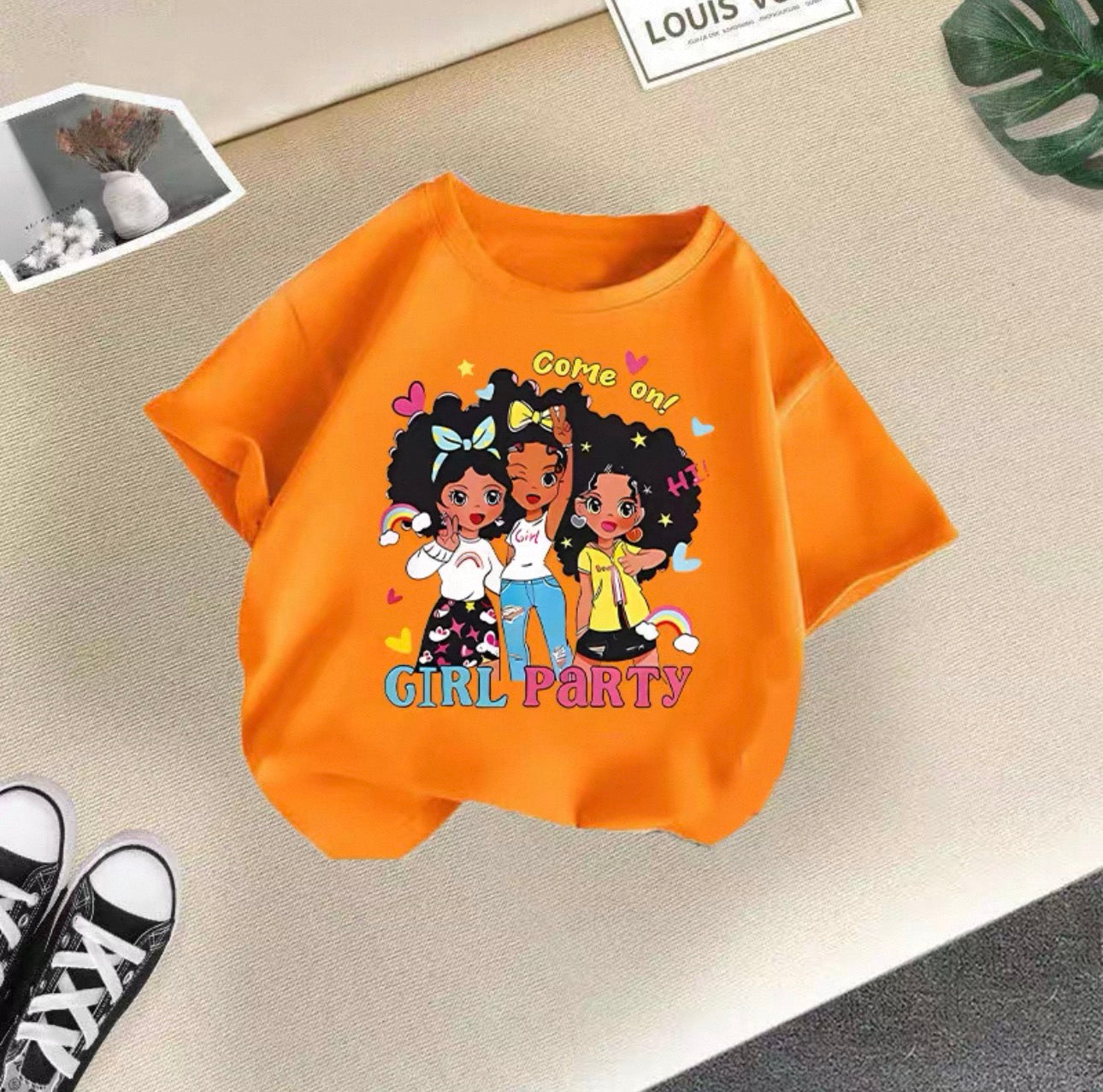 Orange t-shirt with cartoon girls and 'Girl Party' text on a beige surface.