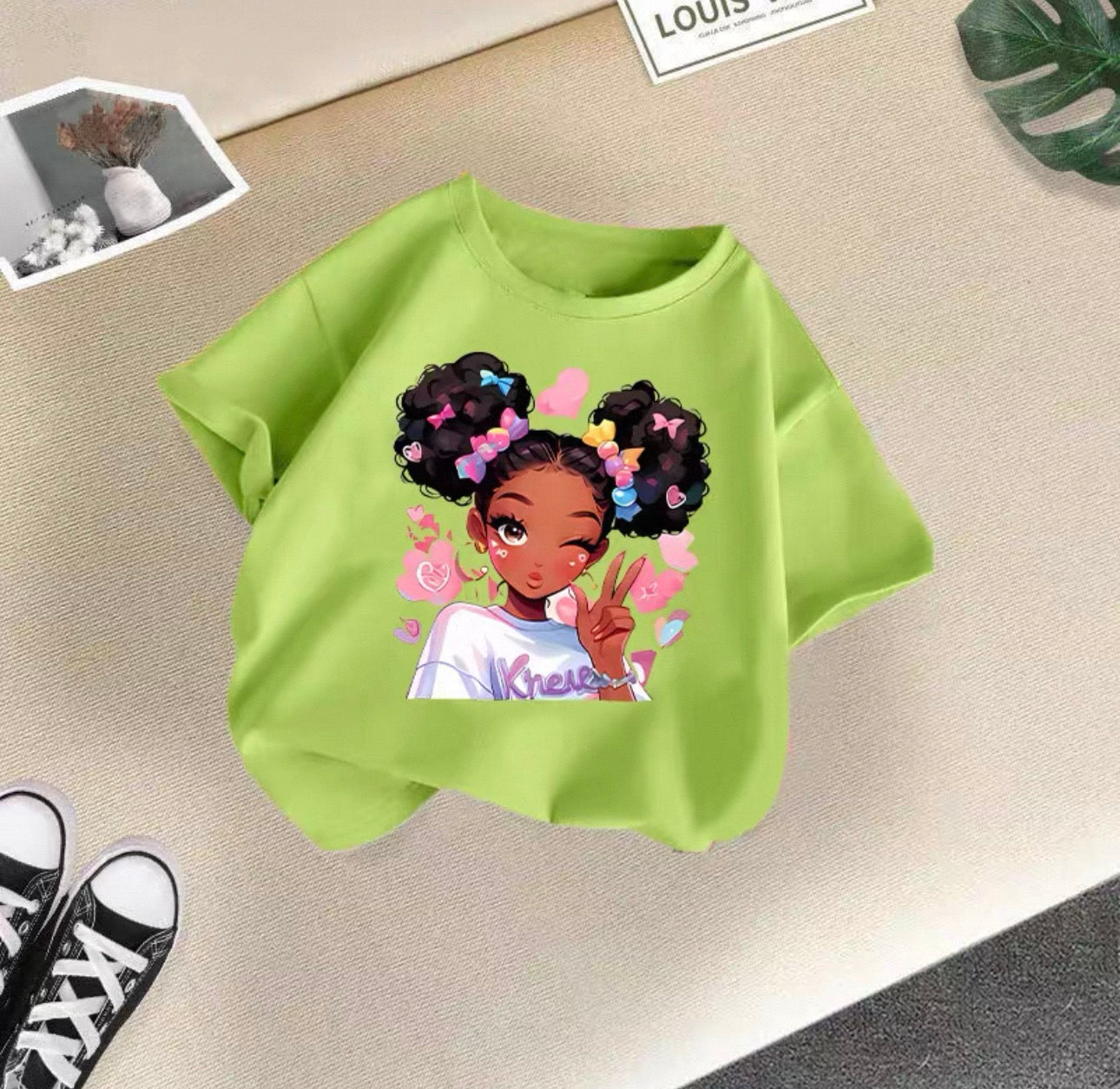 Green t-shirt with a graphic of a girl with braids and flowers on a beige surface.