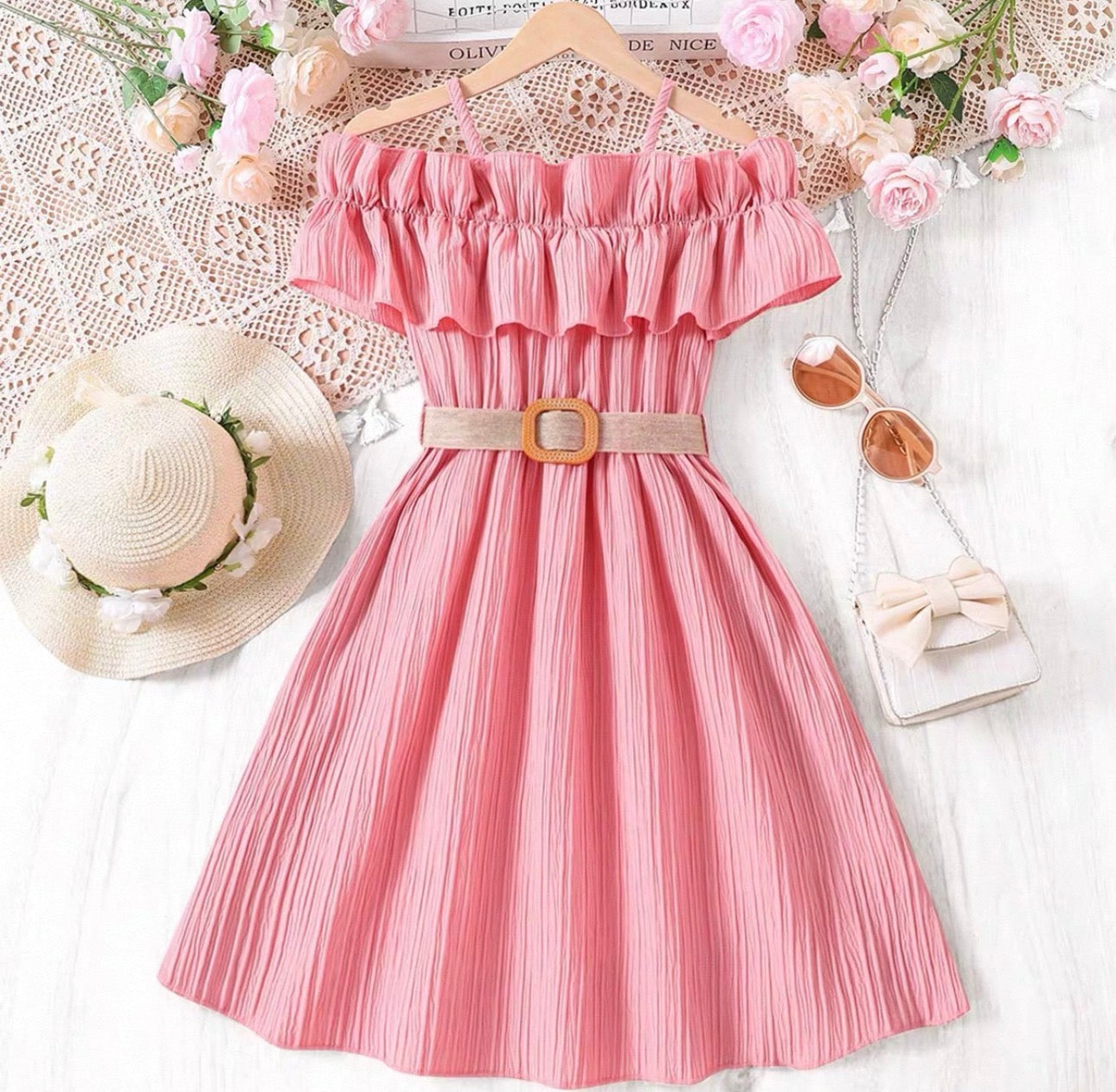 Pink dress with ruffled top and belt, surrounded by flowers and accessories on a white background