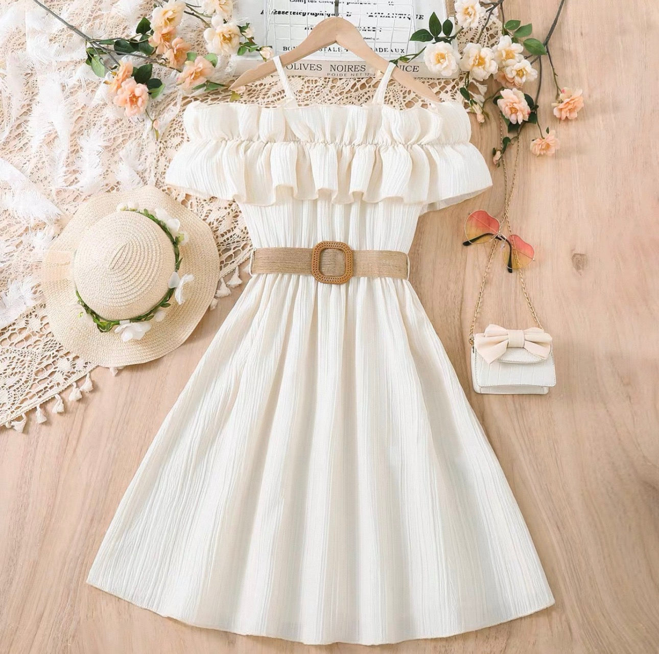 White dress with a belt, straw hat, and small handbag on a wooden surface with flowers.