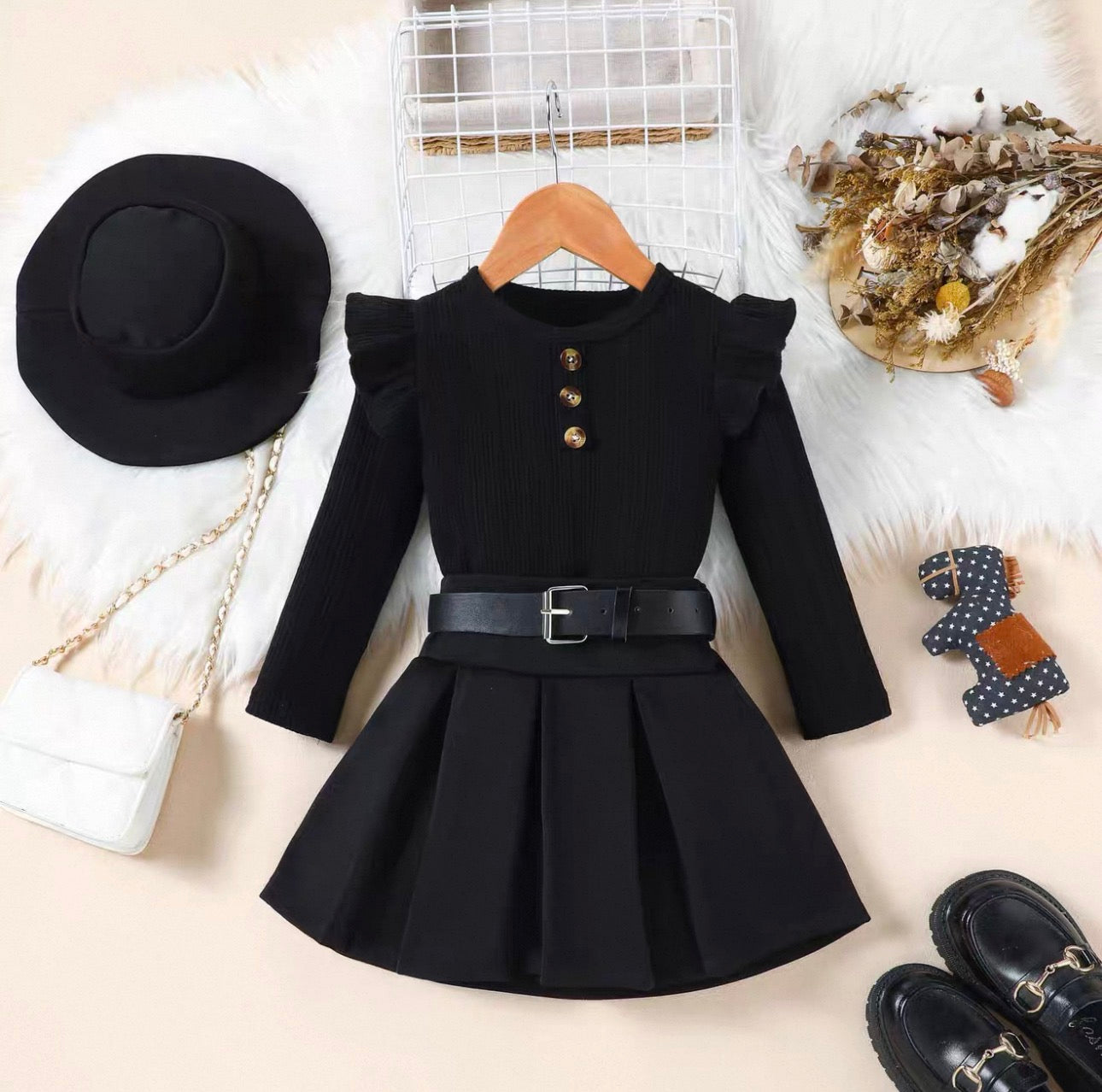 Black dress with a belt on a hanger, surrounded by a black hat, white handbag, and black shoes on a light background.