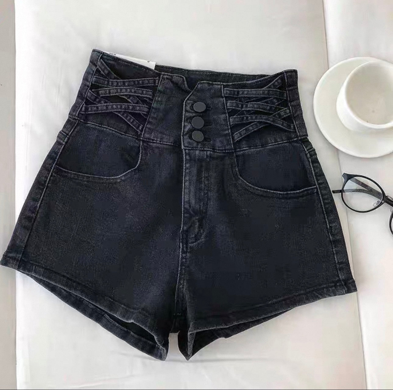 Black denim shorts on a white surface with a cup and glasses in the background