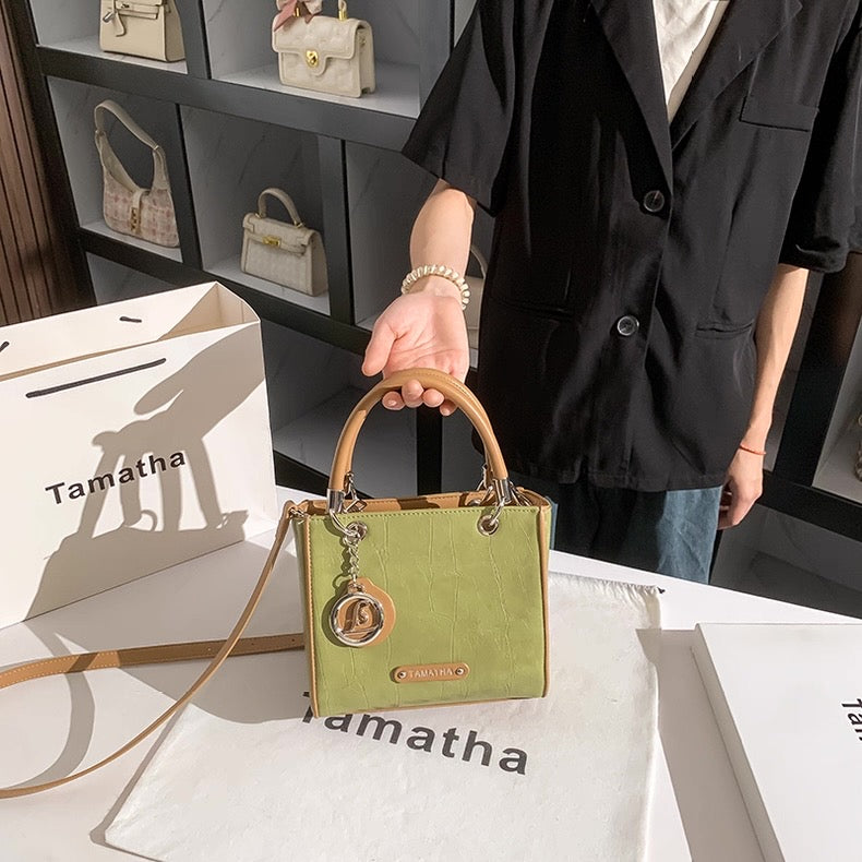 Green handbag with brown handle and strap, displayed on a white surface with 'Tamatha' branding.